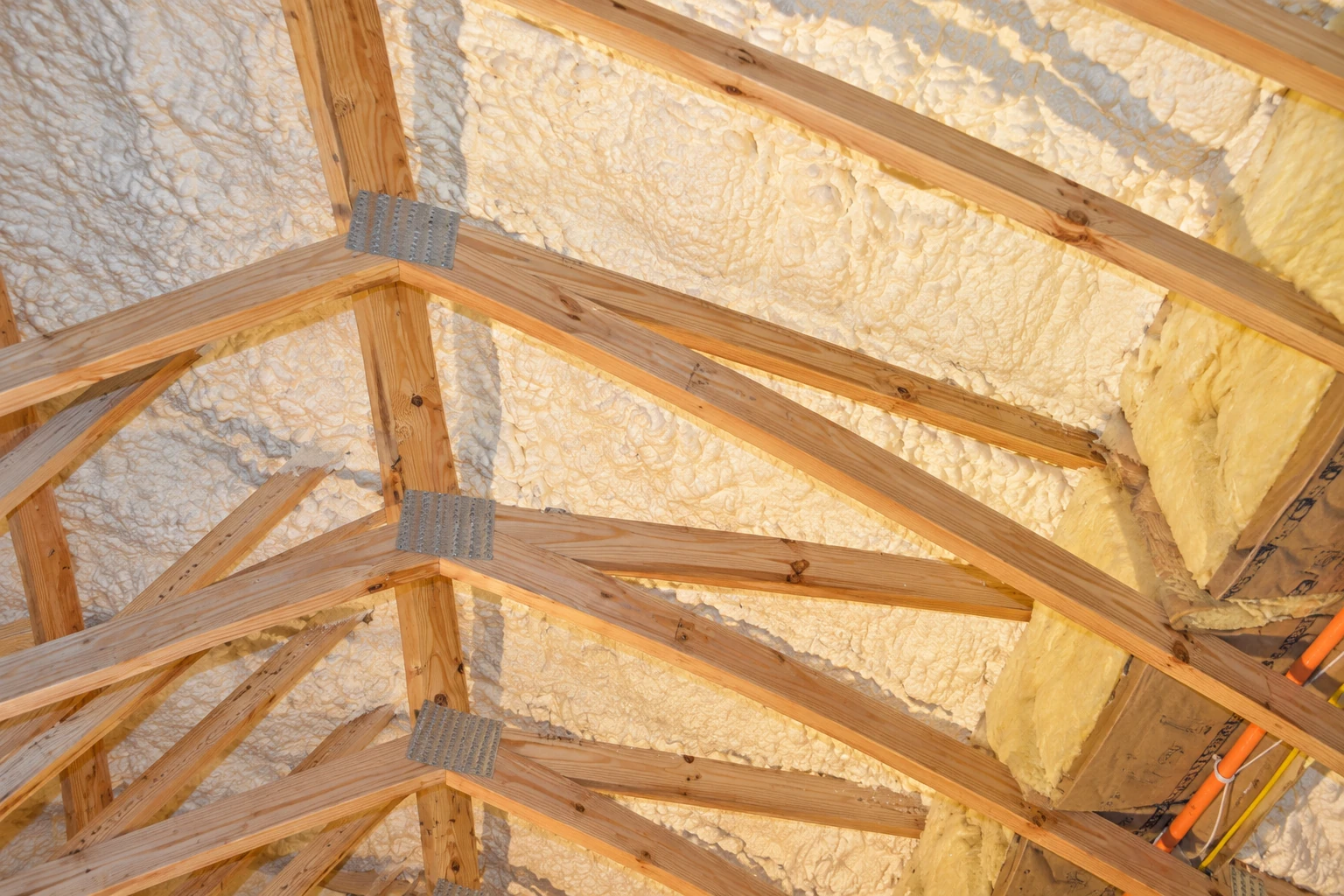 White spray foam covers the roof sheathing between exposed wooden trusses in an attic.
