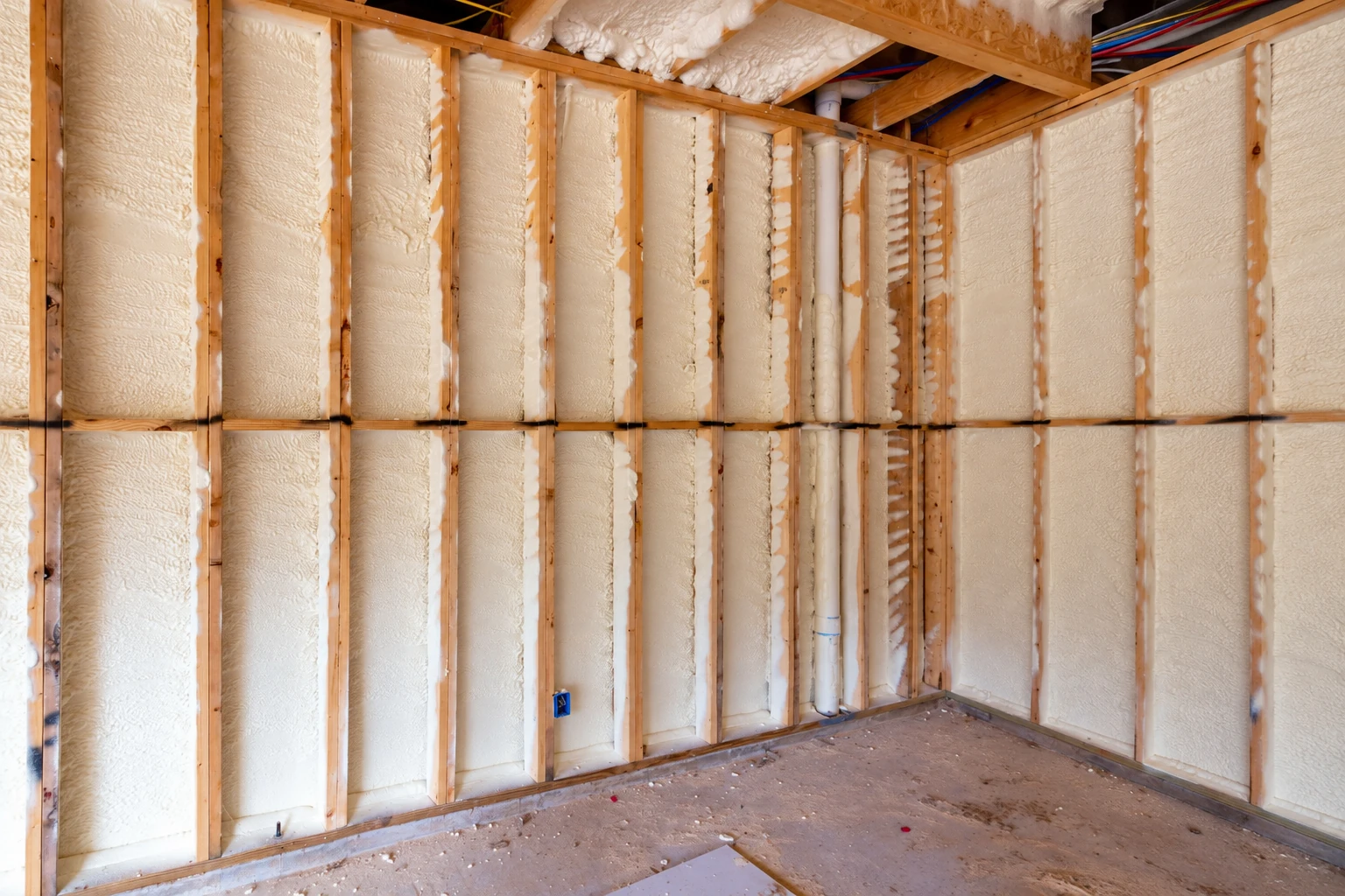 Foam insulation fills the wall cavities between exposed wooden studs in an unfinished room.