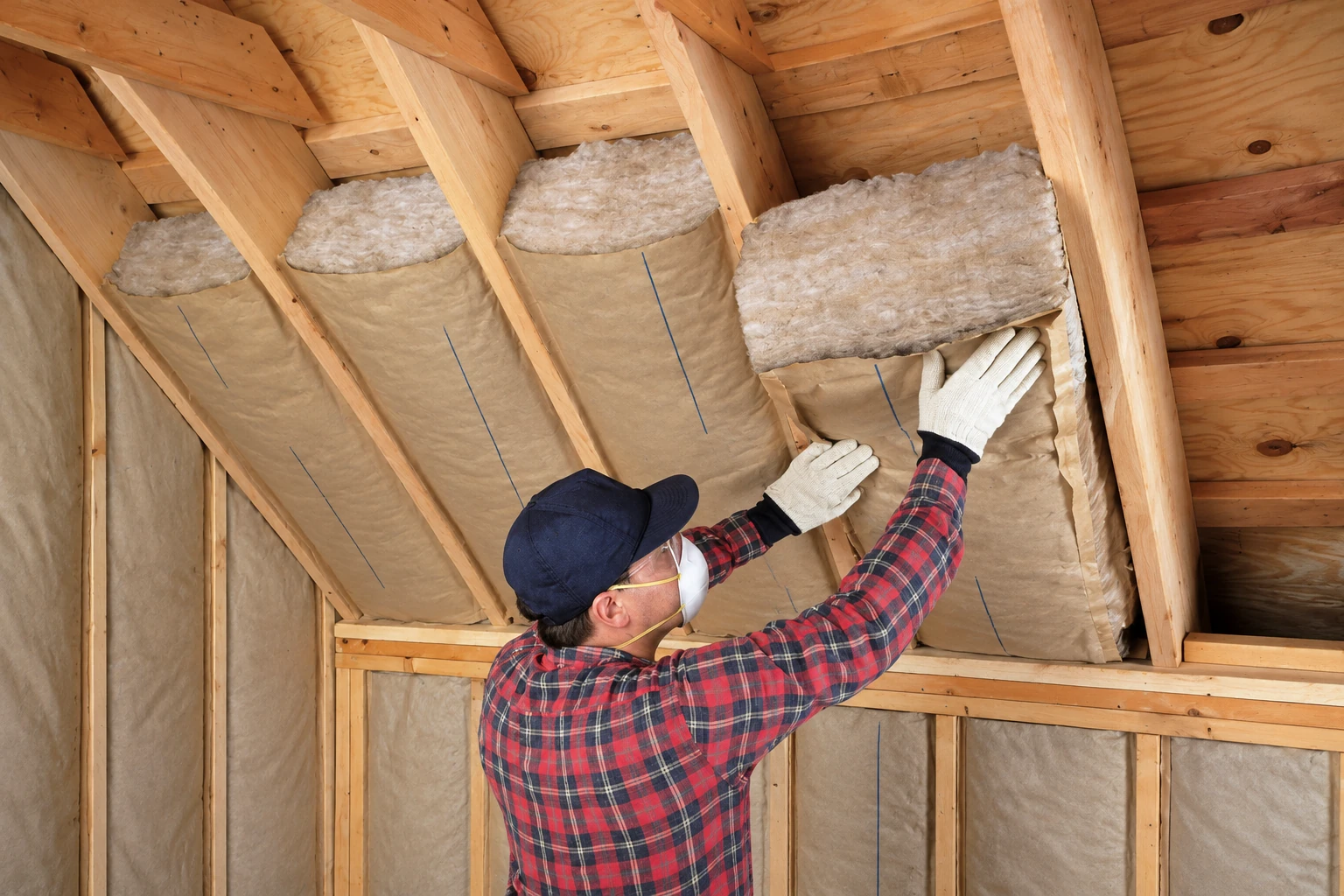 A worker in gloves fits a fiberglass batt between exposed roof rafters.